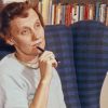 Die Schriftstellerin Astrid Lindgren hat kurzes, rotes, gelocktes Haar. Sie sitzt in einem blauen Sessel vor einem Bücherregal und blickt nachdenklich vor sich hin. Dabei hält sie einen Kugelschreiber an ihr Kinn.