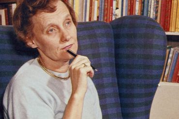 Die Schriftstellerin Astrid Lindgren hat kurzes, rotes, gelocktes Haar. Sie sitzt in einem blauen Sessel vor einem Bücherregal und blickt nachdenklich vor sich hin. Dabei hält sie einen Kugelschreiber an ihr Kinn.