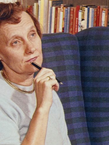Die Schriftstellerin Astrid Lindgren hat kurzes, rotes, gelocktes Haar. Sie sitzt in einem blauen Sessel vor einem Bücherregal und blickt nachdenklich vor sich hin. Dabei hält sie einen Kugelschreiber an ihr Kinn.