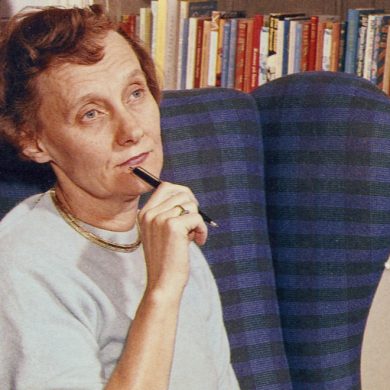 Die Schriftstellerin Astrid Lindgren hat kurzes, rotes, gelocktes Haar. Sie sitzt in einem blauen Sessel vor einem Bücherregal und blickt nachdenklich vor sich hin. Dabei hält sie einen Kugelschreiber an ihr Kinn.