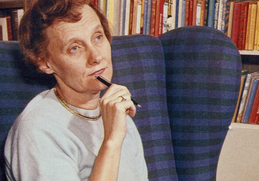 Die Schriftstellerin Astrid Lindgren hat kurzes, rotes, gelocktes Haar. Sie sitzt in einem blauen Sessel vor einem Bücherregal und blickt nachdenklich vor sich hin. Dabei hält sie einen Kugelschreiber an ihr Kinn.
