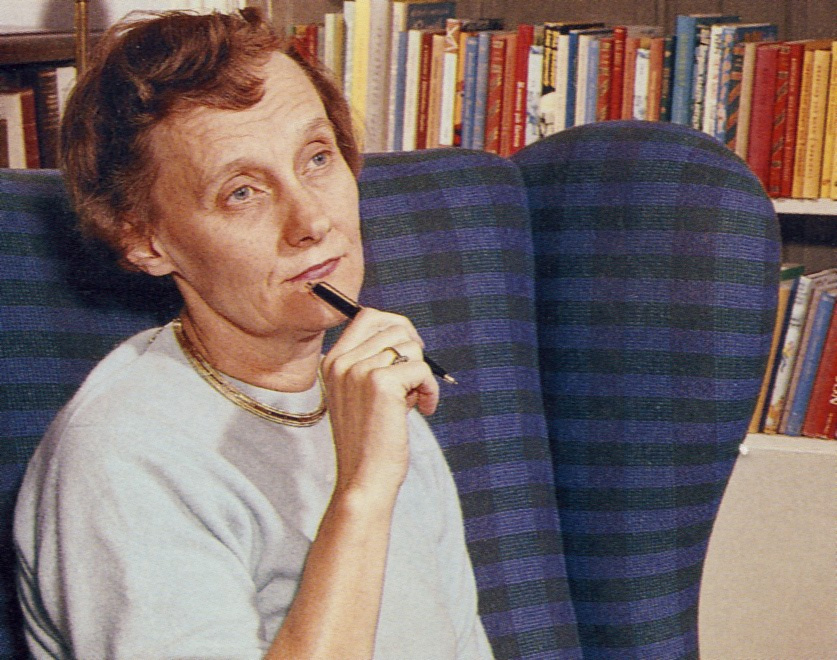 Die Schriftstellerin Astrid Lindgren hat kurzes, rotes, gelocktes Haar. Sie sitzt in einem blauen Sessel vor einem Bücherregal und blickt nachdenklich vor sich hin. Dabei hält sie einen Kugelschreiber an ihr Kinn.