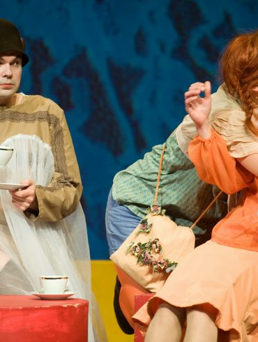 Das Bild zeigt drei Personen, die auf der Bühne sind: Sitzend eine Person mit Hut, die eine Teetasse hält, und eine weitere Person mit Handtasche, die eine Bluse und einen Rock trägt. Die dritte Person ist als Pippi Langstrumpf verkleidet und küsst die Person mit der Handtasche auf die Wange.