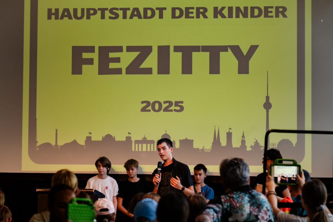 Kinder stehen auf der Bühne: Die Kinderregierung stellt sich vor - auf der FEZitty-Eröffnung am 30. Juli 2025. Hinter den Kindern ist das FEZitty-Stadtzeichen zu sehen. Es zeigt die Skyline Berlins auf gelbem Grund. Darauf geschrieben steht: "Hauptstadt der Kinder FEZitty 2025".