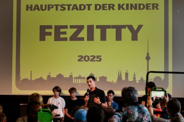 Kinder stehen auf der Bühne: Die Kinderregierung stellt sich vor - auf der FEZitty-Eröffnung am 30. Juli 2025. Hinter den Kindern ist das FEZitty-Stadtzeichen zu sehen. Es zeigt die Skyline Berlins auf gelbem Grund. Darauf geschrieben steht: "Hauptstadt der Kinder FEZitty 2025".