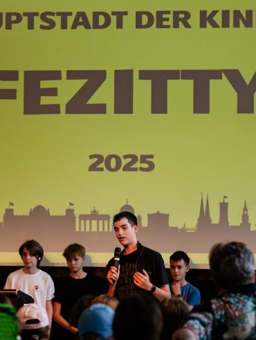 Kinder stehen auf der Bühne: Die Kinderregierung stellt sich vor - auf der FEZitty-Eröffnung am 30. Juli 2025. Hinter den Kindern ist das FEZitty-Stadtzeichen zu sehen. Es zeigt die Skyline Berlins auf gelbem Grund. Darauf geschrieben steht: "Hauptstadt der Kinder FEZitty 2025".