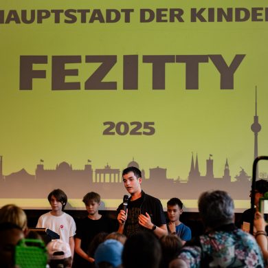 Kinder stehen auf der Bühne: Die Kinderregierung stellt sich vor - auf der FEZitty-Eröffnung am 30. Juli 2025. Hinter den Kindern ist das FEZitty-Stadtzeichen zu sehen. Es zeigt die Skyline Berlins auf gelbem Grund. Darauf geschrieben steht: "Hauptstadt der Kinder FEZitty 2025".