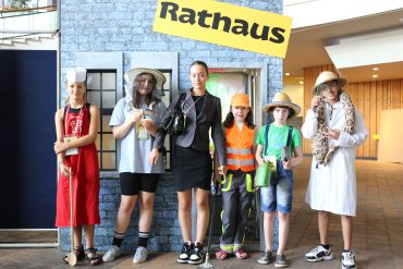 Sechs Kinder stehen vor einem Pappmaché-Haus, auf dem "Rathaus" geschrieben steht.