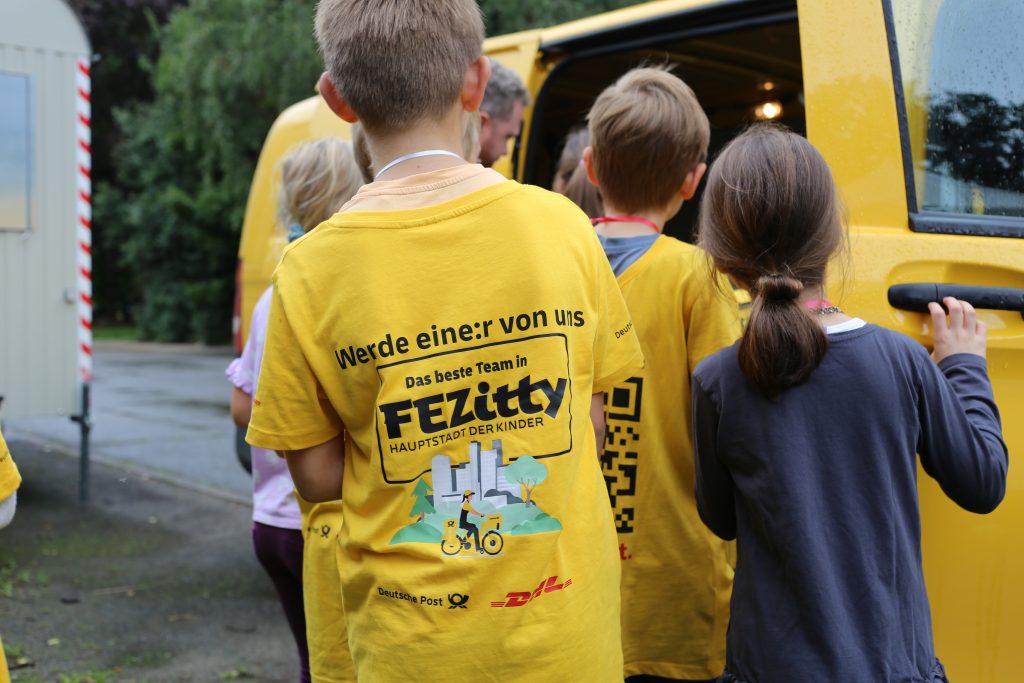 Eine Gruppe von Kindern begutachtet ein elektrisches Zustellfahrzeug der Deutschen Post. Die meisten von ihnen tragen gelbe FEZitty-T-Shirts.